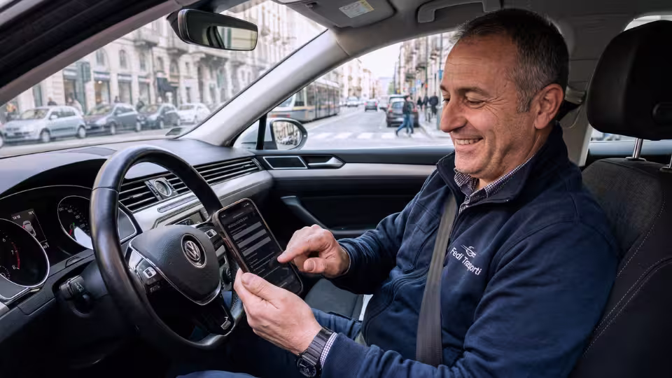 A candid photograph taken inside a car shows a man in a blue jacket smiling while looking at and interacting with a smartphone held in his hand.