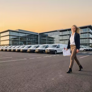 A businesswoman smiling and walking in a corporate parking lot with a long row of commercial vans. In the background, a modern glass office building with 'FUSION SOLUTIONS' branding stands under a golden-hour sunset sky. The image represents the integration of advanced VIN-recognition and AI-operations in corporate fleet management.